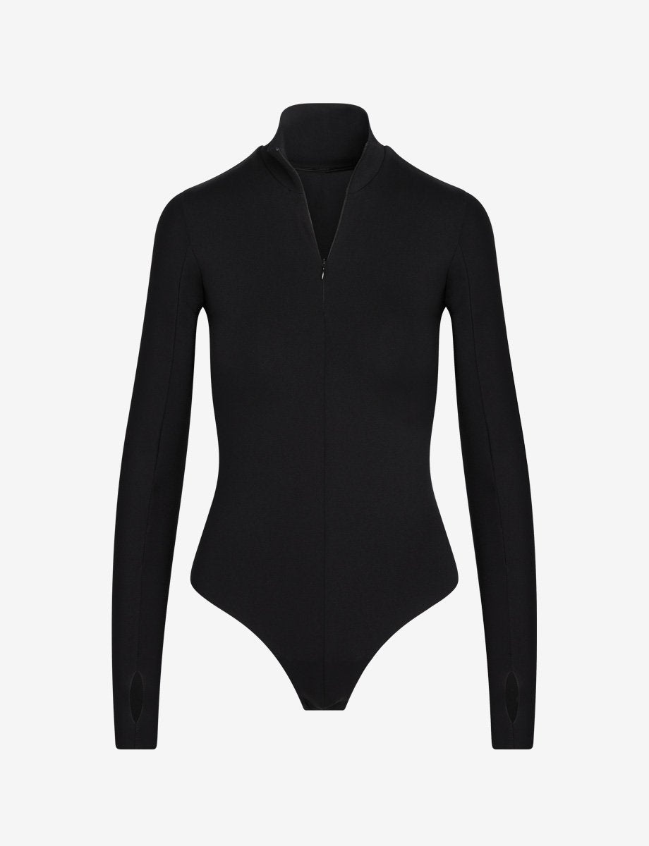 UNIKONCEPT Shoppe & Lounge: The model is wearing the Neoprene Zip Long Sleeve Bodysuit by Commando. This black bodysuit has thumb holes and features raw cut edges. It has a quarter zip at the neckline with a collar and has a strap gusset closure. It is made from stretchy neoprene and hugs curves perfectly.
