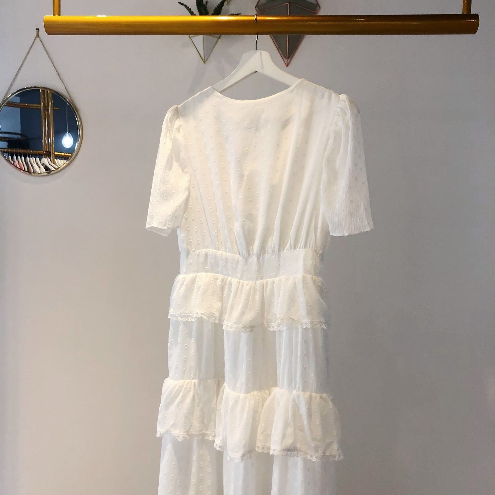 UNIKONCEPT Lifestyle boutique: The image shows the Hamptons White Dress by Free the Roses. This white, ankle length button down dress is extremely elegant with a v-neck and relaxed billowing short sleeves. The fabric is made with small white dots on the white base to add some dimension. The long skirt is tiered and ruffled with a lattice lace detailing as a trim on each layer. 